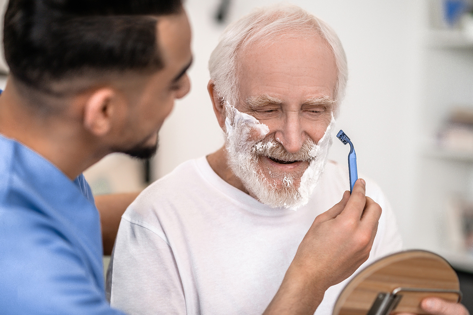 Man sieht einen Pfleger, wie er einen älteren Mann im Gesicht rasiert.