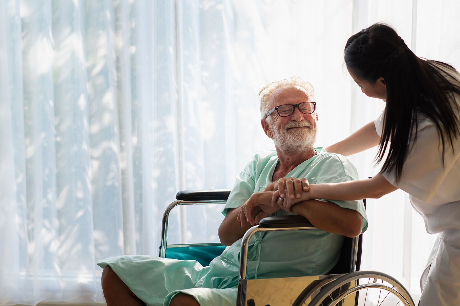 Ein ölterer Mann istzt im Rollstuhl. Eine Pflegerin reicht ihm seine Hand. Er lächelt sie an.