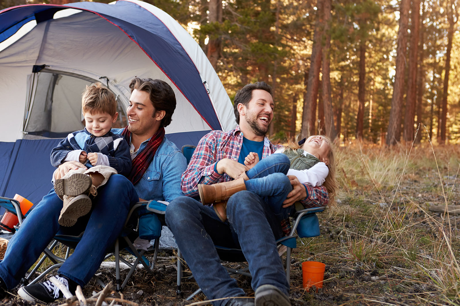 Zwei Männer sitzten mit Kindern vor einem Zelt beim Camping. Alle lachen.
